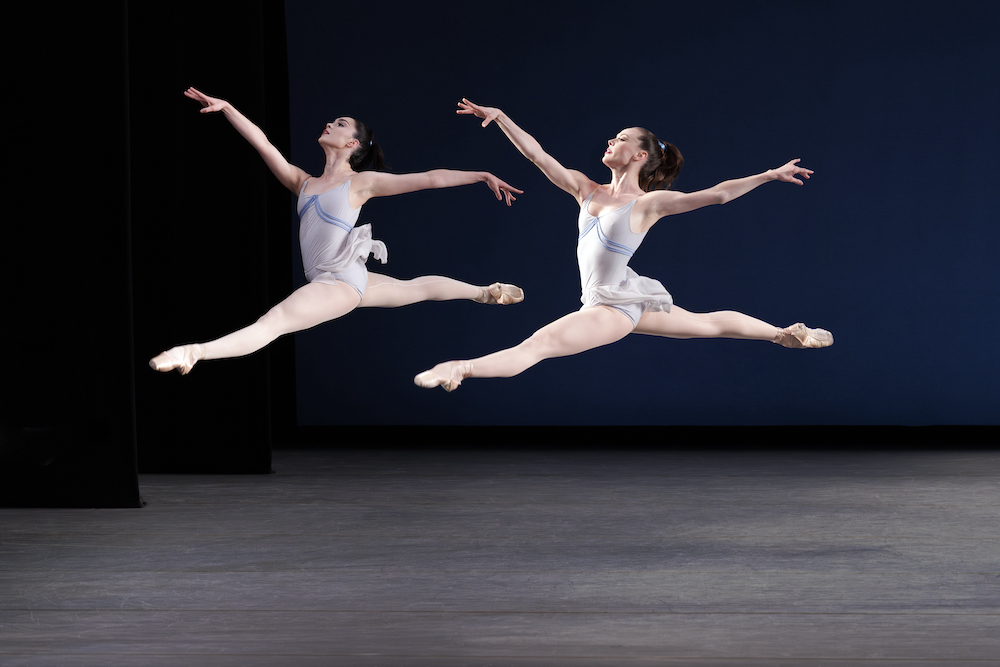 two ballerinas in light blue leotards with short flowing skirts mirror each other in grande jetes or big jumps 